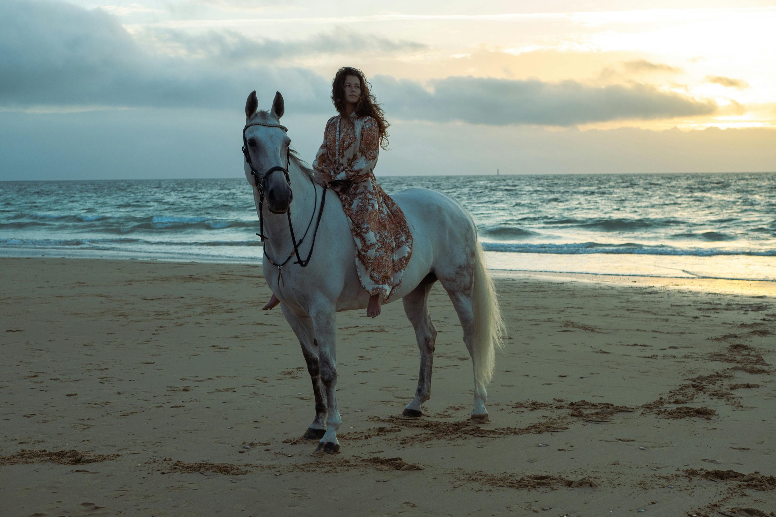 Woman in sundress rides a horse on a serene beach in Nouvelle-Aquitaine at sunset.